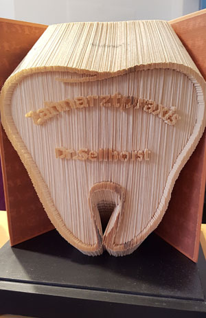 Eine Skulptur in Form eines Zahns, gefertigt aus Holz, mit dem eingravierten Text 'Zahnarztpraxis Dr. Seibhorst'. Die Skulptur steht auf einem schwarzen Sockel.